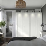Modernes Schlafzimmer mit weißem PAX-Kleiderschrank, grauen Wänden, dunklem Boden und Lampe. Blaues Bett im Vordergrund, Badezimmertür offen.