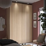 Holzschrank mit Schiebetüren, Stuhl und Kissen. Bett mit Hemd, Fotos und Pflanze am Fenster.