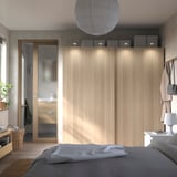 Modernes Schlafzimmer mit PAX-Kleiderschrank aus Eiche, Schiebetüren, integrierter Beleuchtung. Graues Bett mit weißer Decke, Topfpflanze, Korb und Hängetaschen.