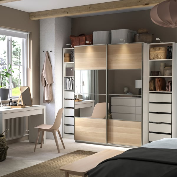 Modernes Schlafzimmer mit PAX-Kleiderschrank. Helles Holz, verspiegelte Türen, Regale, Schubladen. Schreibtisch, Stuhl, Pflanzen, neutrale Töne.