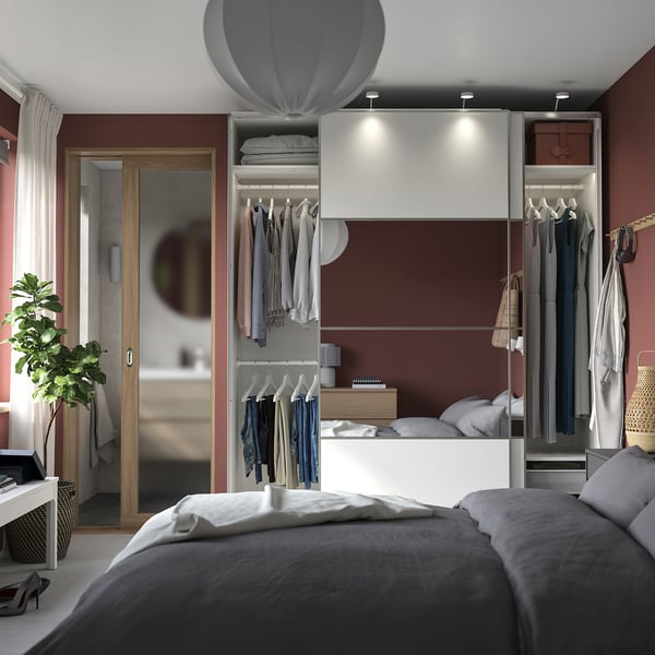 Modernes Schlafzimmer mit weißem PAX-Kleiderschrank, Kleiderbügeln, Spiegel, grauem Bett, roter Wand und Topfpflanze.