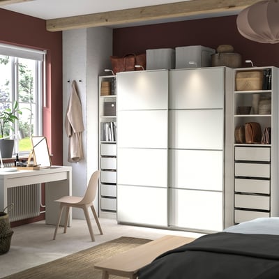 Modernes Schlafzimmer mit einem weißen PAX-Kleiderschrank mit Schiebetüren und Regalen. Enthält einen Schreibtisch, einen Stuhl und natürliche Dekorationselemente.