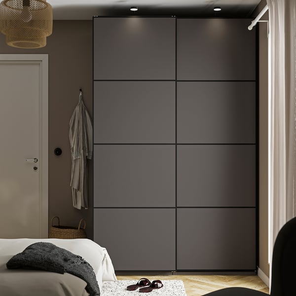 Modernes Schlafzimmer mit einem großen, schlanken dunkelgrauen PAX-Kleiderschrank, der die Wand ausfüllt, einer angebauten Tür mit hängender Jacke und einem ordentlich gemachten Bett darunter.