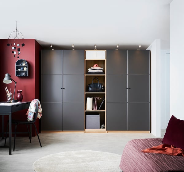 Modernes Schlafzimmer mit großem schwarzen PAX-Kleiderschrank, offenen Regalen mit gefalteter Kleidung und Accessoires. Schreibtisch mit Stuhl, rote Akzente.