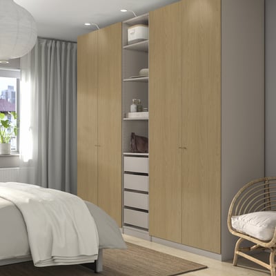 Geräumiges Schlafzimmer mit PAX-Kleiderschrank aus hellem Holz, offenem Bereich, weißem Teppich, Korbstuhl, Fenster, grauen Vorhängen und Bett mit weißer Bettwäsche.