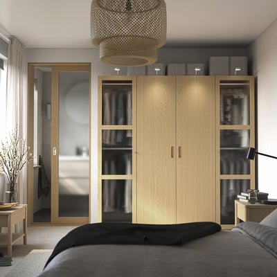 Ruhiges Schlafzimmer mit hohem PAX-Kleiderschrank, Glasscheiben, grauen Boxen, schwarzem Bett, Seitenlampe und warmem Tageslicht.
