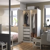Ein modernes Schlafzimmer mit einem weißen PAX-Kleiderschrank, der offen ist, um Kleiderbügel, Schubladen und Aufbewahrungsboxen zu zeigen. Das Zimmer verfügt über ein Bett, Fenster, eine Pflanze und einen Sessel.