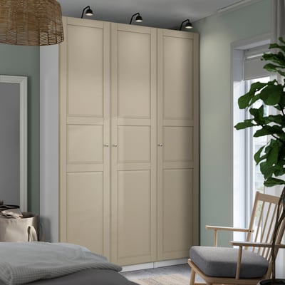 Großer beiger PAX-Kleiderschrank mit Soft-Close-Scharnieren, der in der Nähe eines Fensters mit grüner Pflanze, Stuhl und Bett positioniert ist.