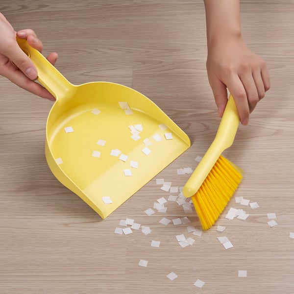 Person mit gelber Kehrschaufel und Bürste, die kleine weiße Stücke vom hellen Holzboden reinigt.