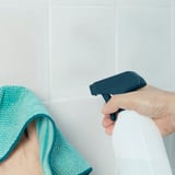 Bildbeschreibung: Eine Hand hält eine PEPPRIG-Sprühflasche und sprüht Reiniger auf eine weiß geflieste Fläche. Ein grünes Tuch liegt in der Nähe.