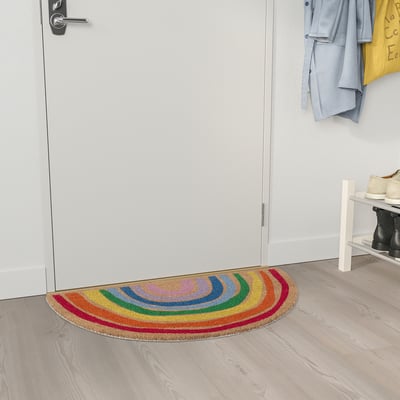 Helle, regenbogenfarbene, halbkreisförmige Fußmatte aus Kokos in der Nähe einer weißen Tür. Es ist kleiner als die Tür und hat ein lebendiges, mehrfarbiges Design.