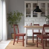 Roter hölzerner PINNTORP Stuhl mit schräger Rückenlehne, um Esstisch auf Teppich, weißer Schrank mit Glaswaren, Pflanze. Gemütlich, minimalistisch, rustikal-modern.