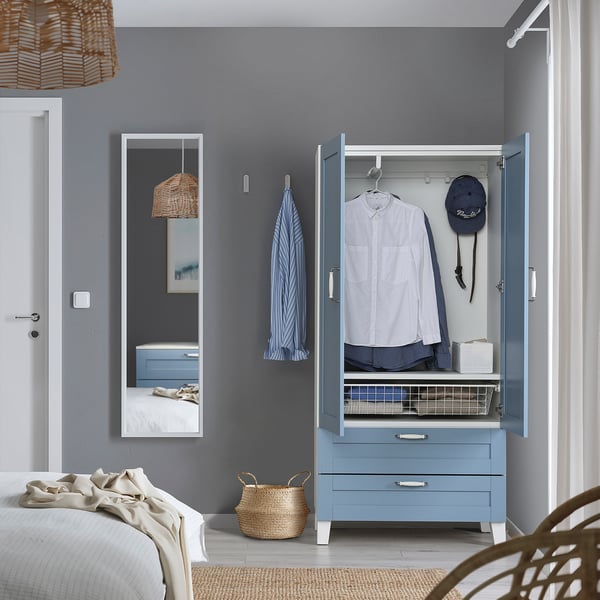 Schlafzimmer mit blauem PLATSA-Kleiderschrank, Kleidung und Schubladen. Weißes Bett, Korb, Handtuch und Spiegel.