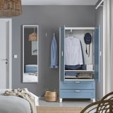 Schlafzimmer mit blauem PLATSA-Kleiderschrank, Kleidung und Schubladen. Weißes Bett, Korb, Handtuch und Spiegel.