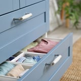 Blaue Kommodenschublade offen, in der ordentlich gefaltete Kleidung in Organizern zu sehen ist. Weiße Griffe sichtbar.