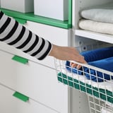 Die Person zieht einen grünen PLATSA-Korb mit gefalteten Handtüchern aus dem weißen Regal.