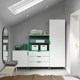Modernes Schlafzimmer mit weißer und grüner PLATSA-Kommode, hohem weißem Schrank, Spiegel und Holzbank unter abgewinkelter Decke.