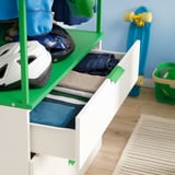 Offene weiße Schublade mit gefalteter Kleidung, daneben Helme auf grünem Regal.