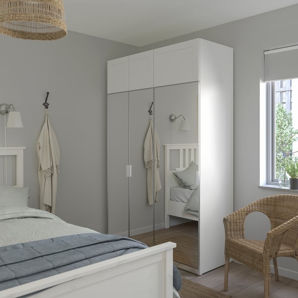 Modernes Schlafzimmer, hoher weißer PLATSA-Kleiderschrank und Bett mit heller Bettwäsche. Korbstuhl- und Spiegeltüren werten den Raum auf.