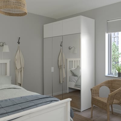 Modernes Schlafzimmer, hoher weißer PLATSA-Kleiderschrank und Bett mit heller Bettwäsche. Korbstuhl- und Spiegeltüren werten den Raum auf.