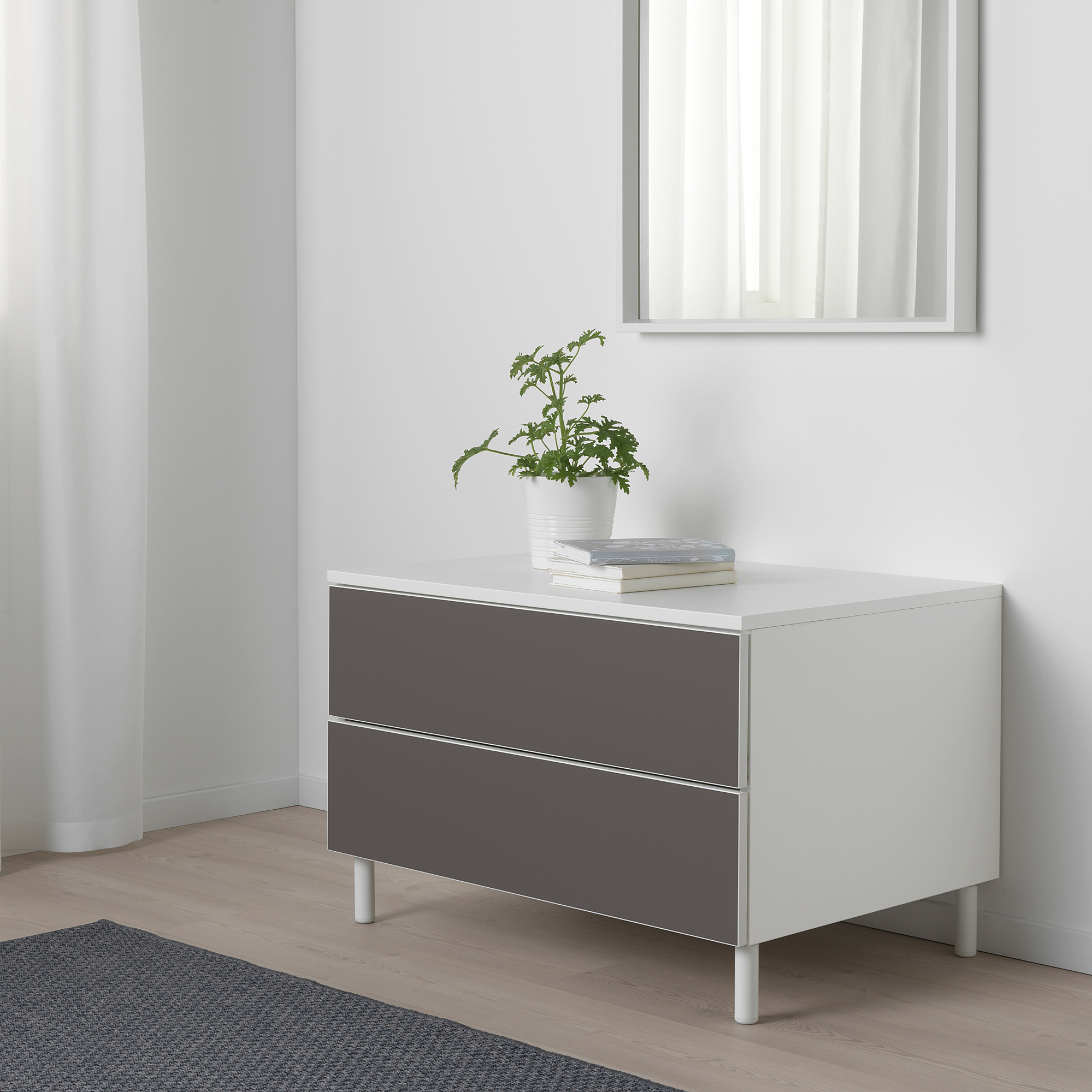 Moderner PLATSA-Stauraum mit 2 grauen Schubladen, weißer Platte, Pflanze, Büchern, vor weißer Wand.