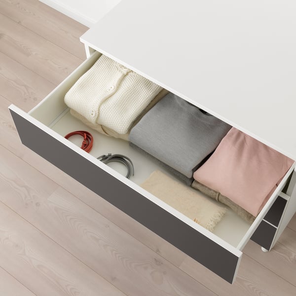 Offene weiße PLATSA-Schublade, die ordentlich gefaltete Kleidung und Kabel auf einem Holzboden freigibt.