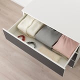 Offene weiße PLATSA-Schublade, die ordentlich gefaltete Kleidung und Kabel auf einem Holzboden freigibt.