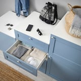 Blauer PLATSA-Stauraum mit offener Schublade mit gefalteter Kleidung und Socken, gekrönt mit Gegenständen wie Jeans, Rucksack und Accessoires.