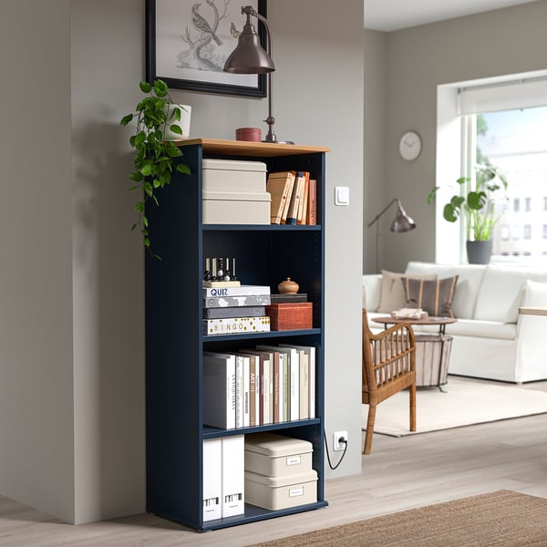 Dunkelblaues PLOGFÅRA Bücherregal mit Büchern, Boxen, Pflanze und Lampe in gemütlichem Wohnraum.