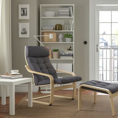 Modernes Wohnzimmer mit grauem POÄNG-Sessel und Fußhocker. Holzrahmen, gepolsterte Sitze, hinter weißem Bücherregal und Fenster.