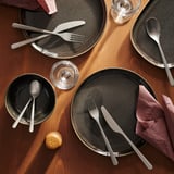 Schwarzes Besteckset auf Tellern, mattes Finish. Messer, Gabel, Löffel. Teller, Gläser, Servietten umgeben.