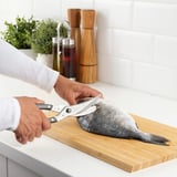 Eine Person schneidet mit einer Schere Fisch auf einem Holzbrett in einer Küche. Die Schere verfügt über einen Verriegelungsmechanismus zur sicheren Aufbewahrung.