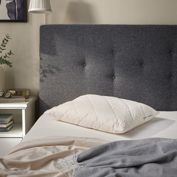 Grau getuftetes Kopfteil mit weißem RÅDHUSVIN-Kissen, heller Bettwäsche, Nachttisch mit Büchern, Lampe und Pflanze.