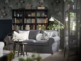 Gemütliches Wohnzimmer mit grauer RANARP Stehleuchte, Sofa, Bücherregalen und dekorativen Pflanzen.