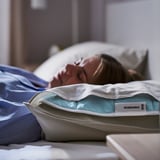 Eine Person, die auf einem Bett ein Nickerchen macht, auf dem ein blaues REXBEGONIA-Kühlkissen liegt. Das Pad hat einen weißen Reißverschluss und ein Etikett.