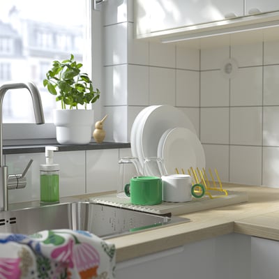 Spülbecken mit RINNIG Abtropffläche, Tellern, Tassen und Besteck. Grünes Spülmittel und Pflanze in der Nähe.