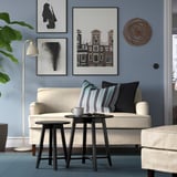 Gemütliches Wohnzimmer mit beigem Sofa, dunklen Kissen, blauer Wand, Kunst, Lampe, Pflanze.