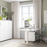 Ein Home-Office-Setup mit einem weißen Sitz-Steh-Schreibtisch RODULF an einem großen Fenster. Ein grauer Stuhl, ein Computer und Pflanzen schmücken den Raum.