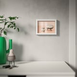 An einer grauen Wand hängt ein moderner weißer Bilderrahmen, in dem minimalistische Möbel in Rosatönen ausgestellt sind. Darunter steht auf einem Schreibtisch eine grüne Vase mit Ästen.