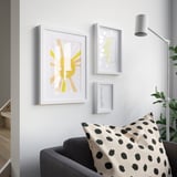 Drei gerahmte Kunstwerke an einer Wand: Sonne, Landkarte, Text. Grauer Sessel mit geflecktem Kissen, Lampe und Pflanze.