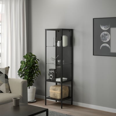Schwarzer RUDSTA-Schrank mit Glastür im modernen Wohnzimmer, in dem eine Vintage-Kamera neben einer großen Pflanze und Mondphasenkunst ausgestellt ist.