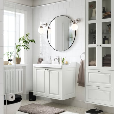 Modernes weißes Badezimmer: runder Spiegel, Wandwaschbecken, Glasvitrine, Pflanzen und Toilettenpapier.