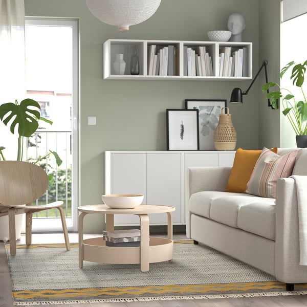 Gemütliches Wohnzimmer mit grünem Ryssgras-Teppich, grauem Sofa, Holztisch und Sessel. Pflanzen drinnen und draußen, dazu ein weißes Regal mit Büchern und Dekoration.
