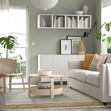 Gemütliches Wohnzimmer mit grünem Ryssgras-Teppich, grauem Sofa, Holztisch und Sessel. Pflanzen drinnen und draußen, dazu ein weißes Regal mit Büchern und Dekoration.