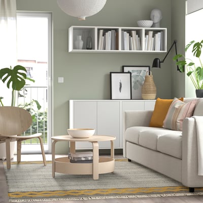 Gemütliches Wohnzimmer mit grünem Ryssgras-Teppich, grauem Sofa, Holztisch und Sessel. Pflanzen drinnen und draußen, dazu ein weißes Regal mit Büchern und Dekoration.