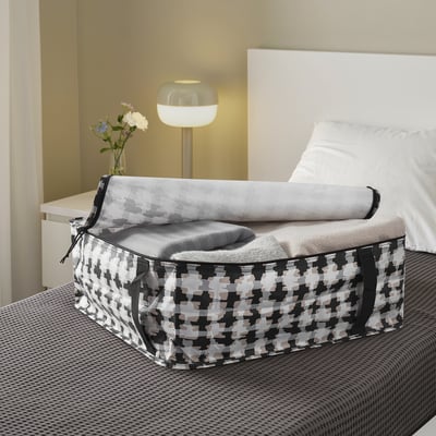 Ein gemusterter Aufbewahrungskoffer auf einem Bett enthält gefaltete Decken, dahinter ein Nachttisch und eine Lampe.