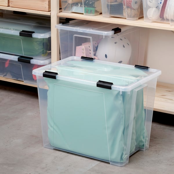 Durchsichtige Aufbewahrungsbox aus Kunststoff, Marke SAMLA, mit schwarzen Clips, die auf dem Boden in der Nähe von Regalen steht. Es ist größer als die Gegenstände um es herum.