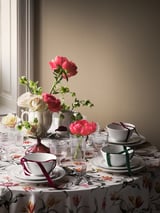 Elegantes Tischset mit floralem Thema. Rosa Rosen, weiße Teetassen mit Bändern und Edelstahlbesteck auf einer geblümten Tischdecke.