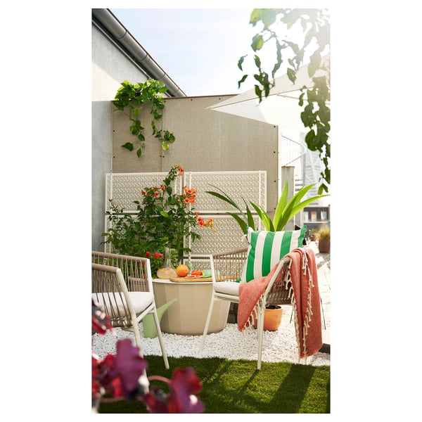 Kleiner Balkon mit weißen Korbstühlen, grünen Kissen und einem Tisch, auf Kunstrasen gestellt, umgeben von Blumen und Pflanzen.