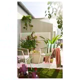 Kleiner Balkon mit weißen Korbstühlen, grünen Kissen und einem Tisch, auf Kunstrasen gestellt, umgeben von Blumen und Pflanzen.
